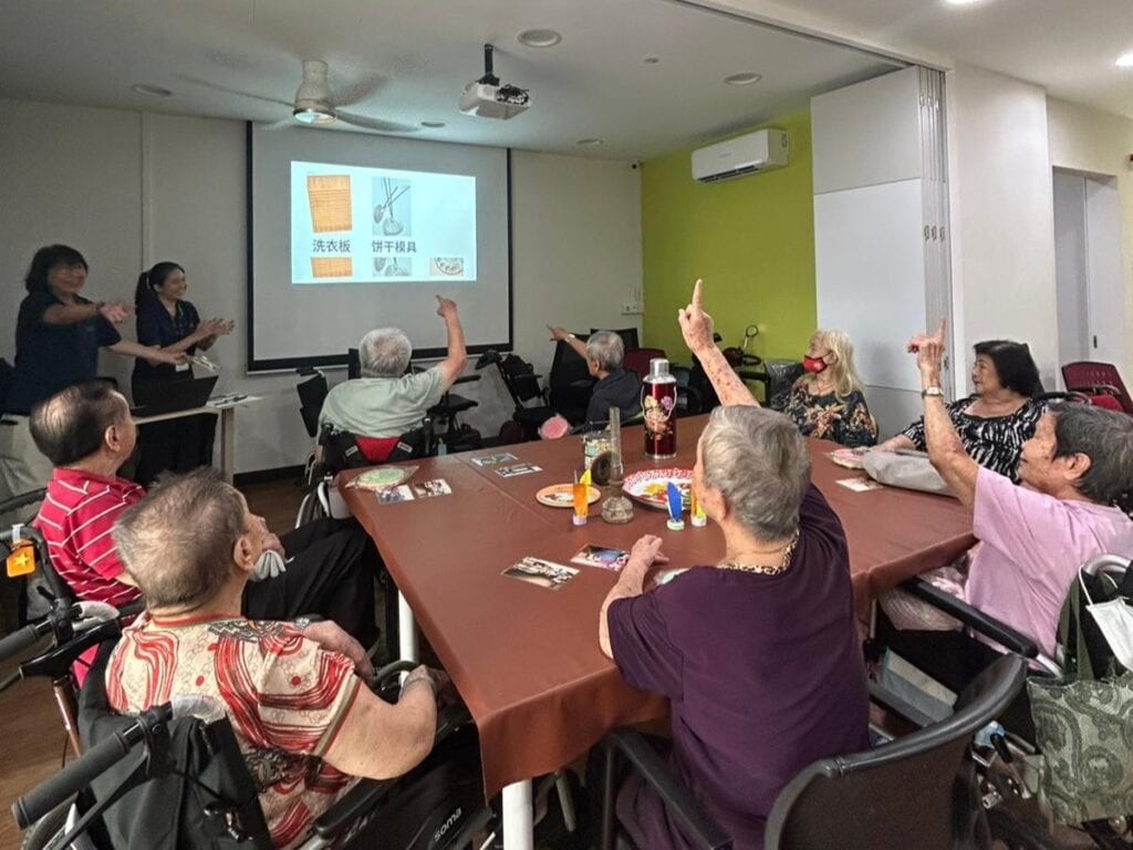 Seniors seated around a square table, one raises her hand to answer about a household item