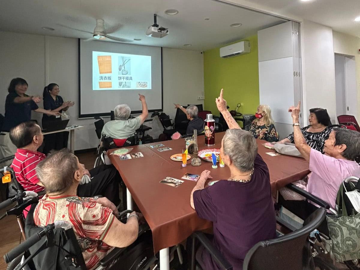 Seniors seated around a square table, one raises her hand to answer about a household item
