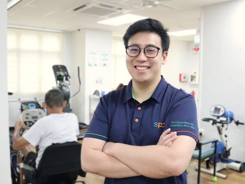 Joel with arms folded, behind him is the rehabilitation centre and a senior exercising