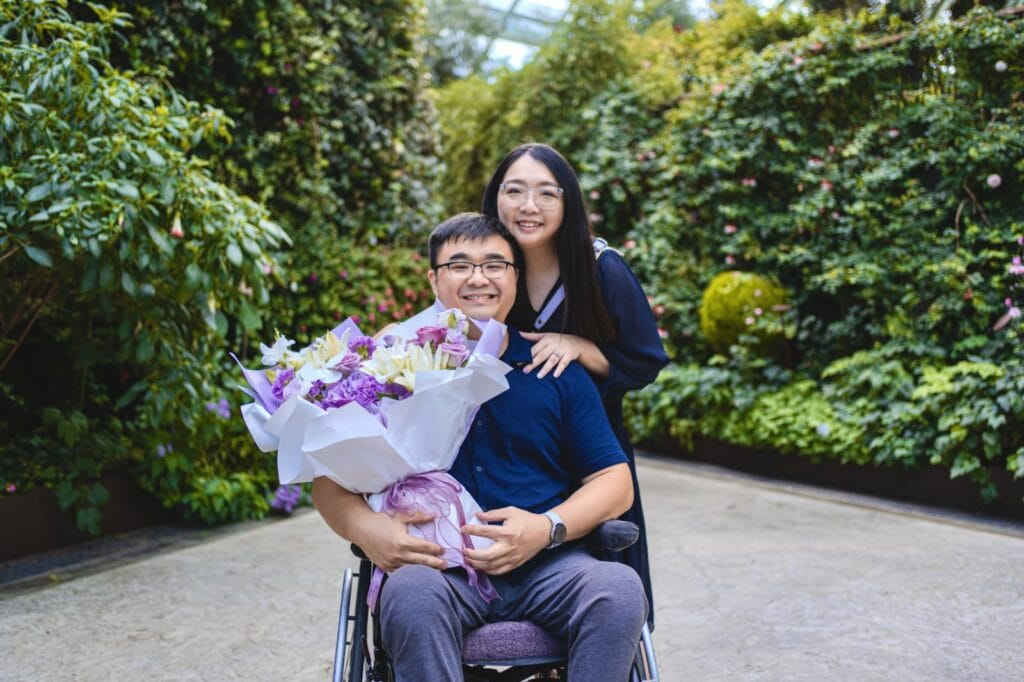 Against a backdrop of trees and greenery, Kelly stands behind Kishon, who is seated in his wheelchair and holding a bouquet of flowers.