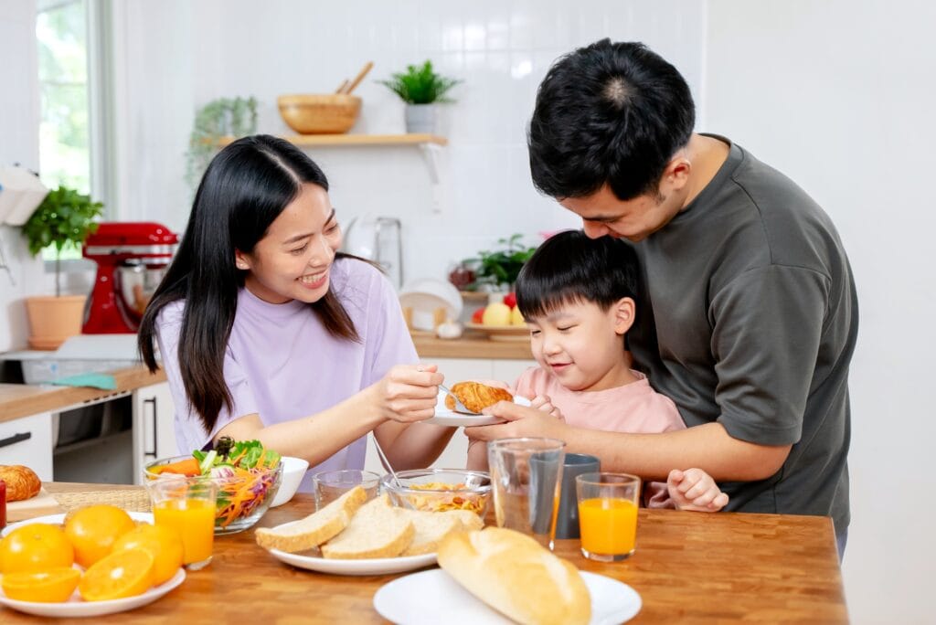 At a meal table with breakfast food like orange juice, salad, cereals and bread, a mother trying to get her child to taste a pastry as he sits on his father's lap.