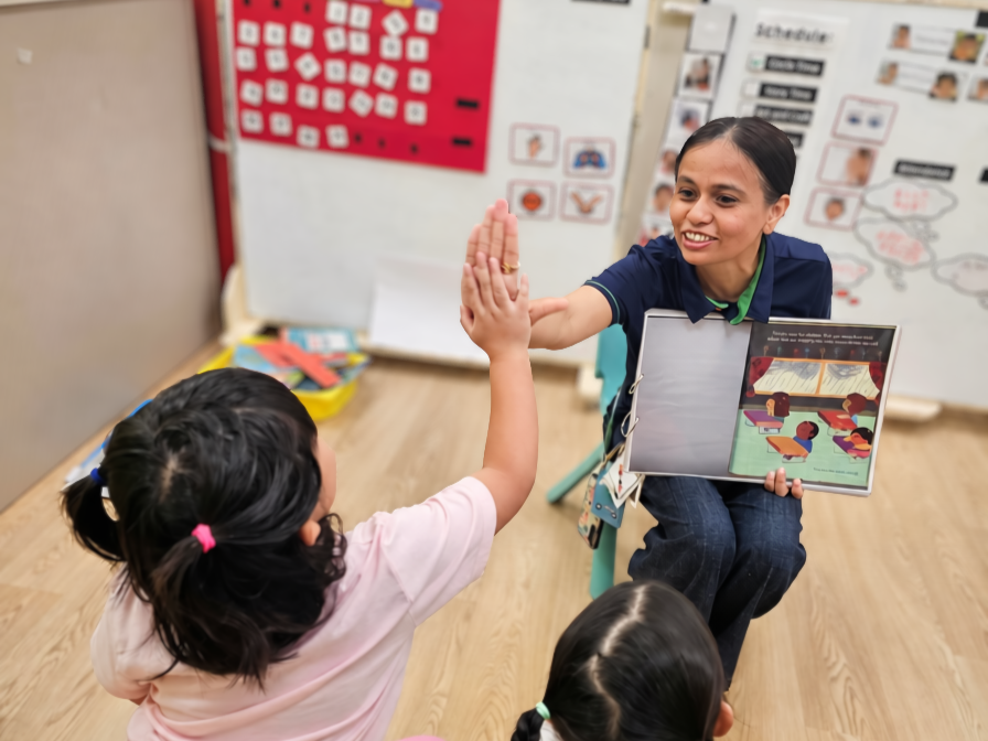 In a classroom, teacher aide Neha is holding a story book and giving a high five to a young child
